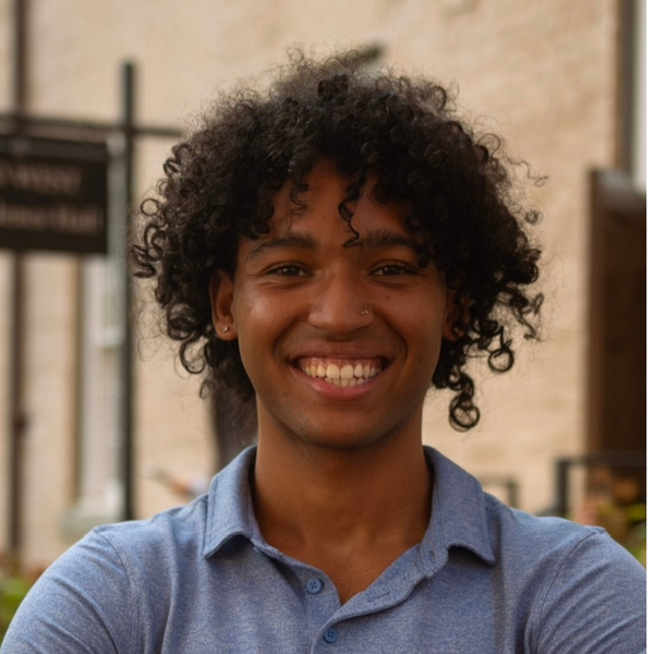A smiling person with curly dark hair and a nose stud stands outdoors with arms crossed, wearing a blue polo shirt against a softly blurred background.